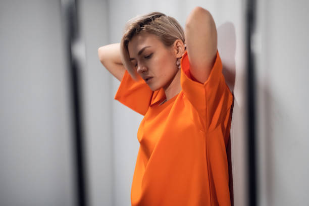 A beautiful blonde criminal in an orange robe stands in her cell behind bars, leaning against the wall.